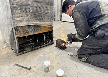 A worker in protective clothing uses an angle grinder on metal equipment outdoors, sparks flying.