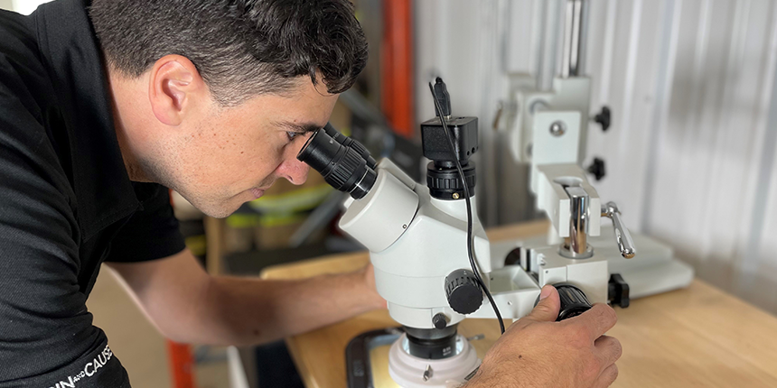 Une personne en chemise noire mène une expérience de microscopie, en ajustant la mise au point sur un microscope blanc placé sur un bureau en bois, avec un autre microscope à l'arrière-plan.