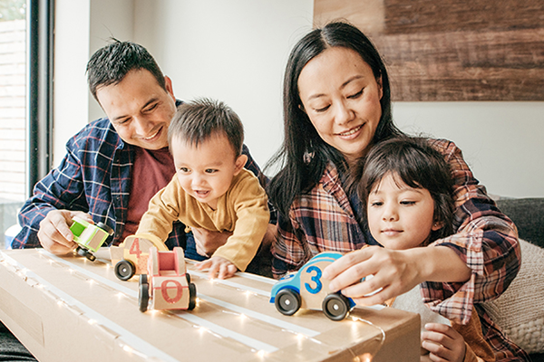 The image shows a family playing with toys indoors. The family consists of a toddler, a baby, and their parents. They are sitting around a table and smiling while sharing the toys.