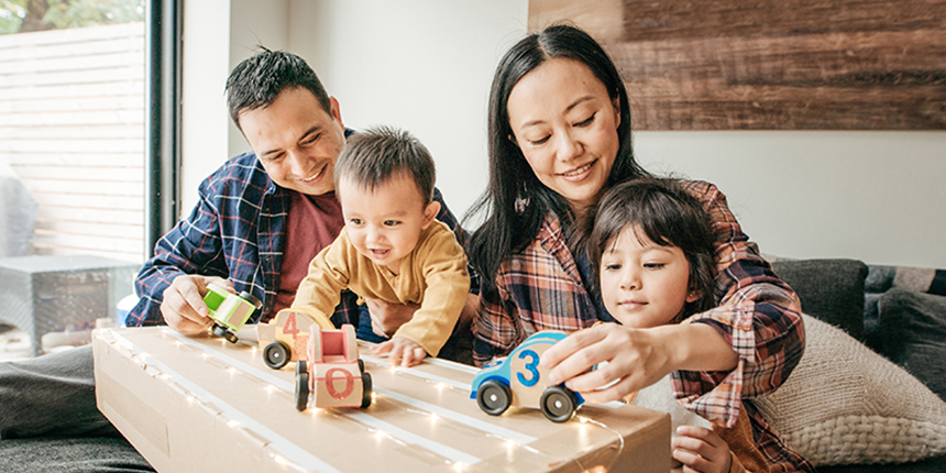 The image shows a family playing with toys indoors. The family consists of a toddler, a baby, and their parents. They are sitting around a table and smiling while sharing the toys.