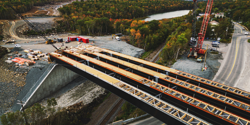 Vue aérienne d'un pont en construction enjambant une route, avec des grues, des matériaux de construction et une rivière entourée d'arbres en arrière-plan.