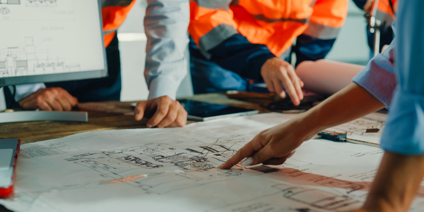 Un groupe de personnes vêtues de gilets de sécurité discutent de plans, en montrant du doigt des dessins architecturaux posés sur une table.